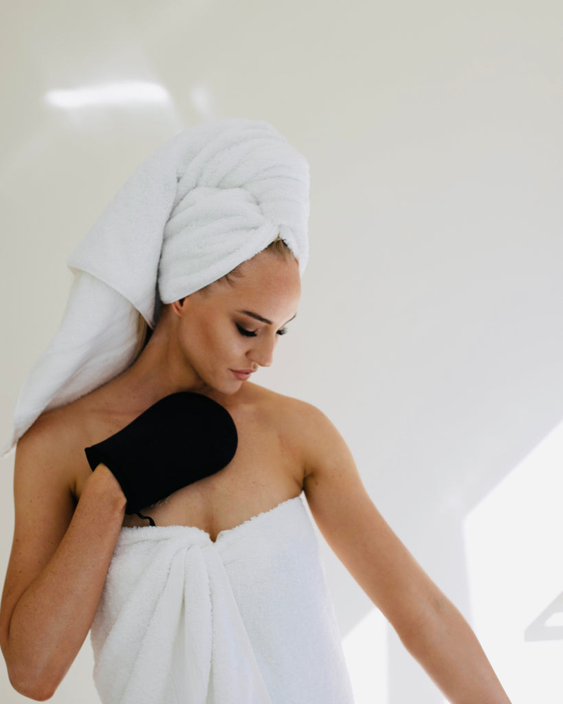 Beautiful tanned woman wearing a white towel with her hair wrapped in a white towel applying fake tan with a black mitt
